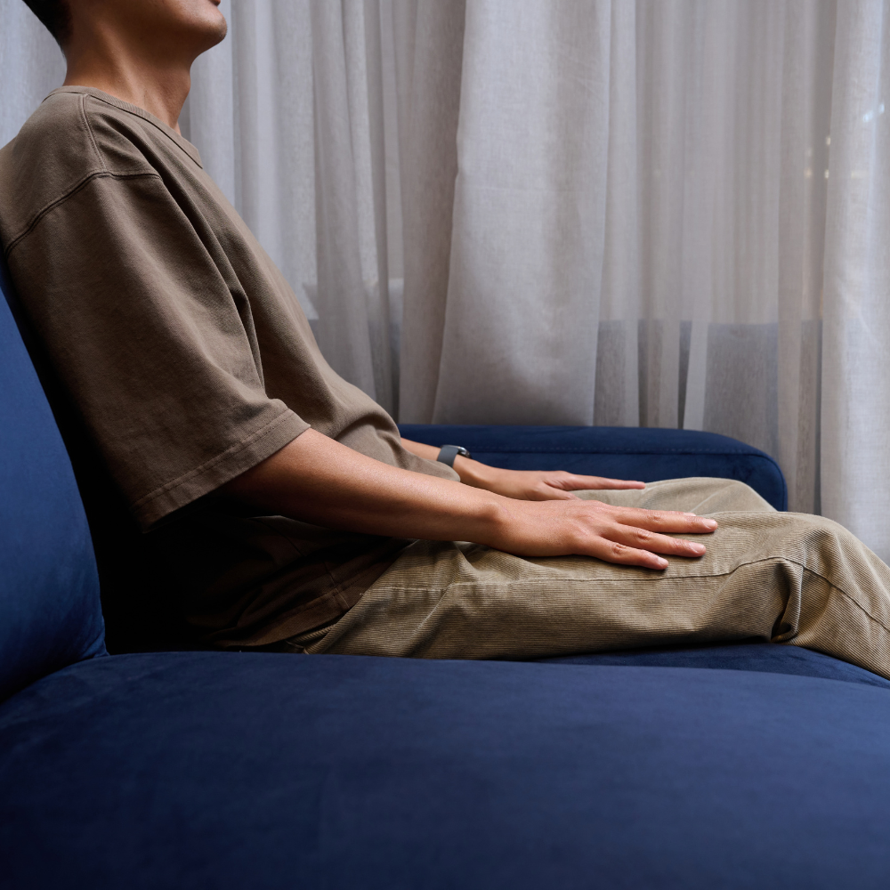 A person sitting on Elise Armchair Navy Blue House Velvet, wearing an oversized taupe-brown short-sleeve t-shirt and light beige trousers, with sheer white curtains in the background.