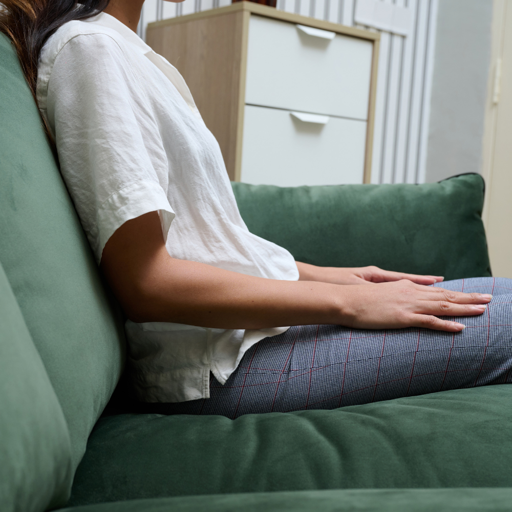 A woman wearing a white short-sleeve button-up shirt and blue plaid trousers sits on ELLE Home Chloe Armchair Moss Green Velvet in a living room setting.