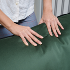 A close-up of a person's hands resting on a ELLE Home Chloe Armchair Moss Green Velvet, showing its smooth texture and sheen. The person is wearing a white button-down shirt and blue plaid pants.